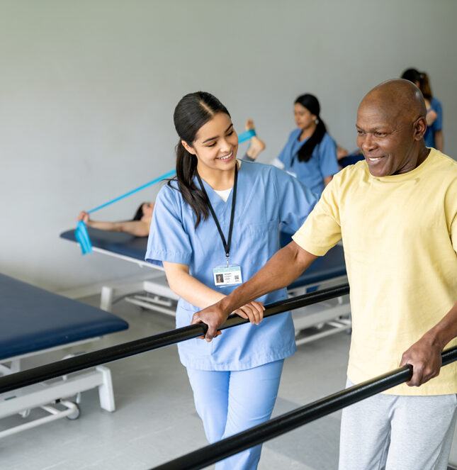 A senior in a gym, while a nurse supports him, participating in orthopedic rehab.